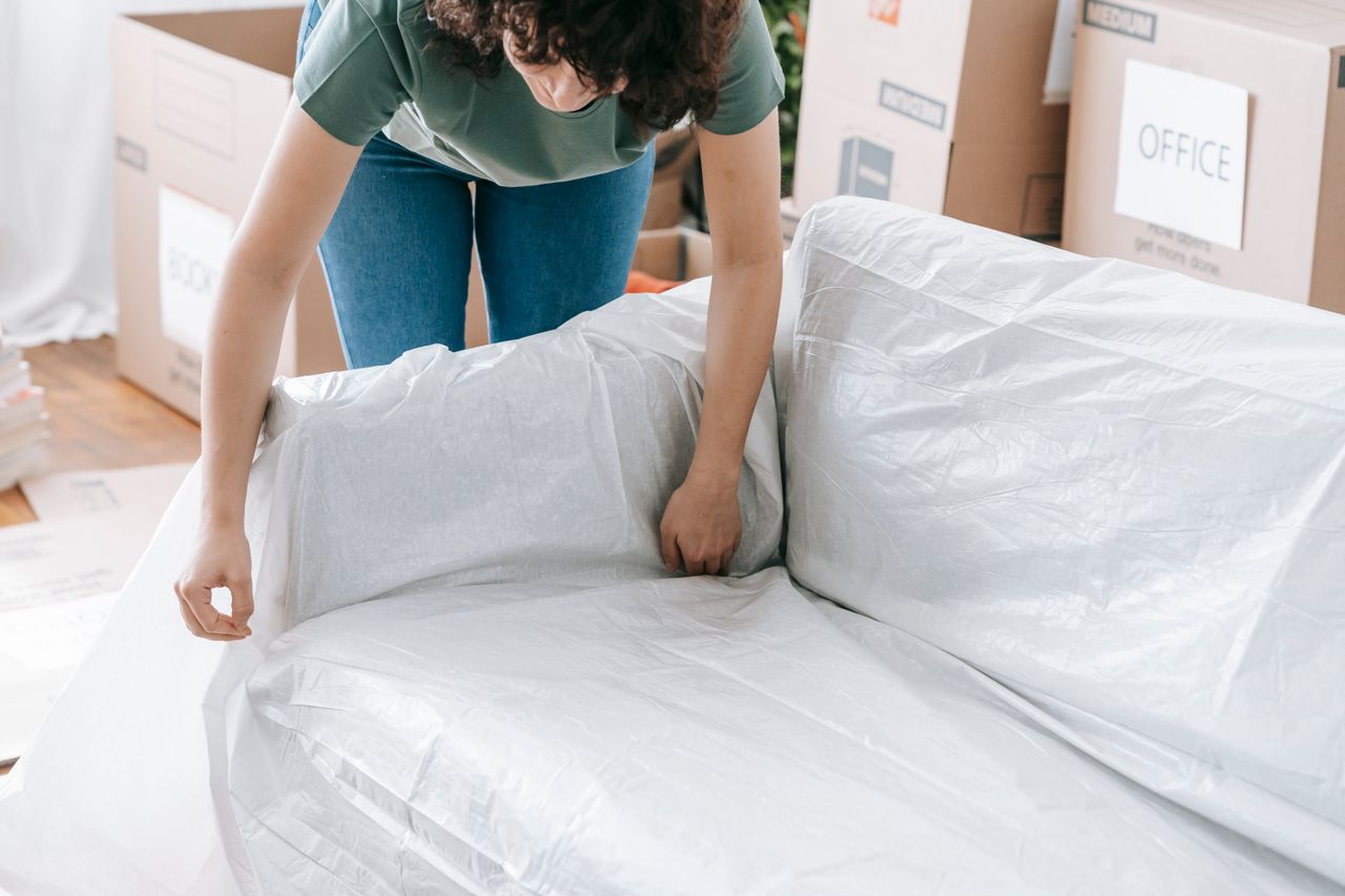A woman meticulously covers a sofa with protective material while packing boxes in a bright room.